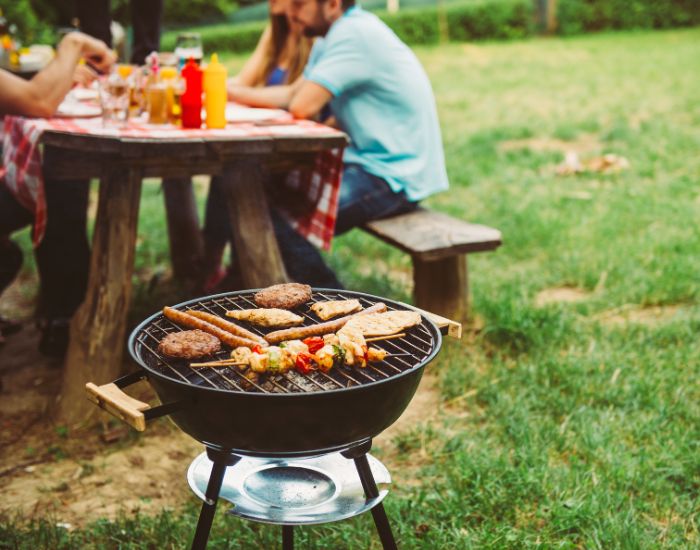 Barbecue grill with chicken, burgers and kebabs cooking in front of people sitting at an outdoor picnic table