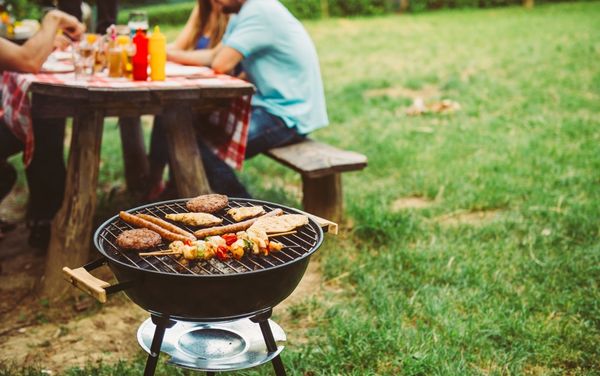 Barbecue grill with chicken, burgers and kebabs cooking in front of people sitting at an outdoor picnic table
