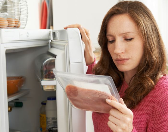 Woman checking the use-by date label on a packet of sliced meat in front of an open fridge