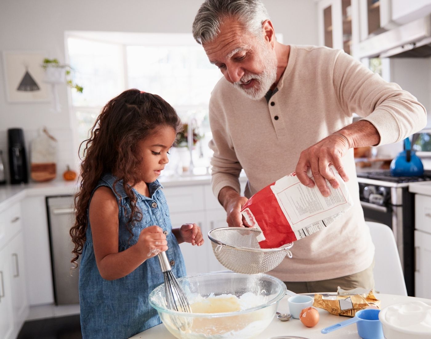 Grandfather baking in the kitchen and sifting flour into a bowl while his granddaughter holds a whisk