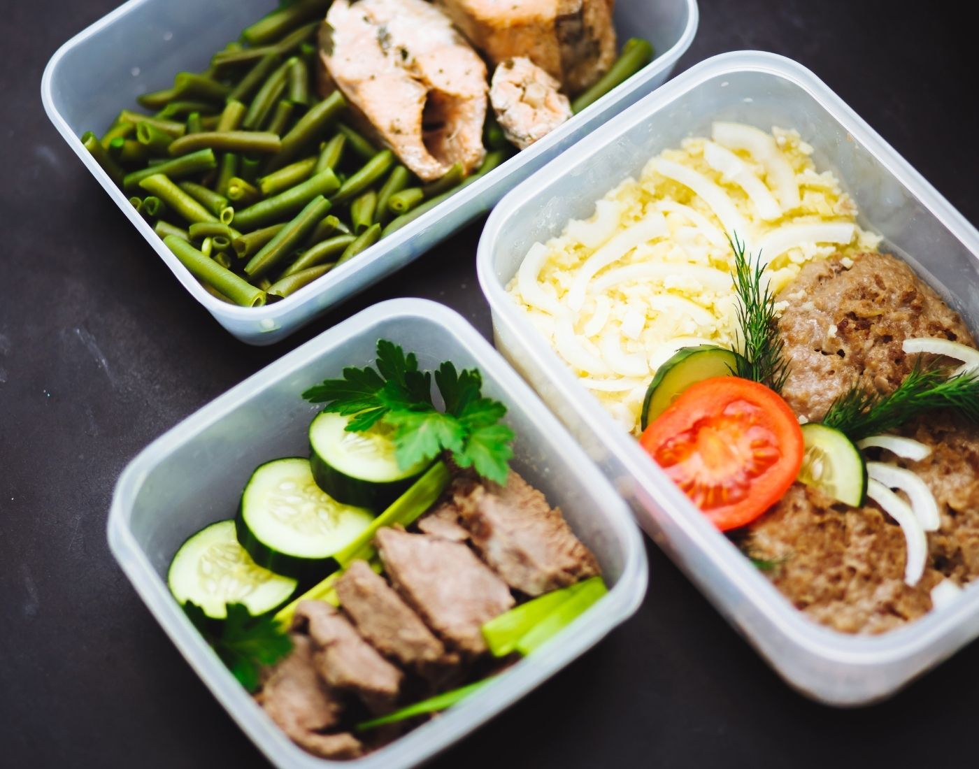 Three plastic food containers with cooked meals including green beans, sliced meat, cucumber and tomato