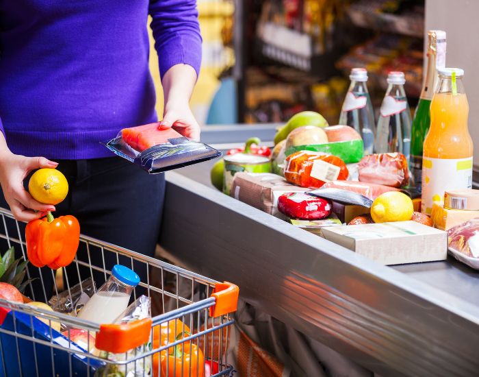 Person loading food items, including fish, fresh produce, and bottled beverages, from a shopping trolley onto a checkout belt in a supermarket