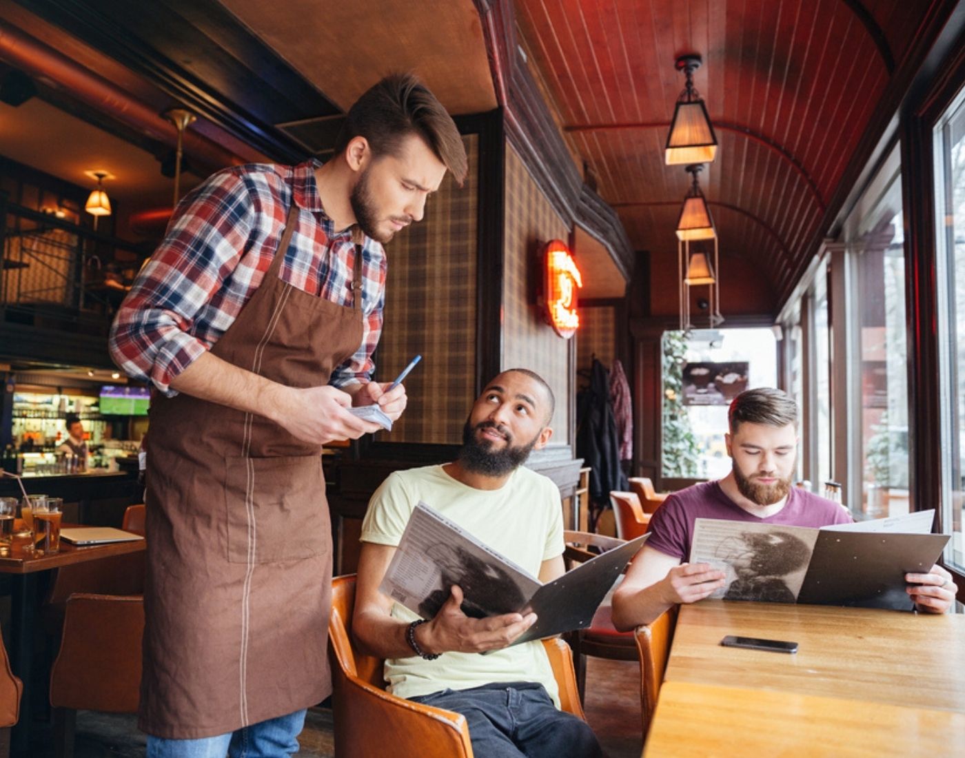 A waiter in an apron taking a food order from a customer seated at a table in a restaurant, while another customer looks at a menu