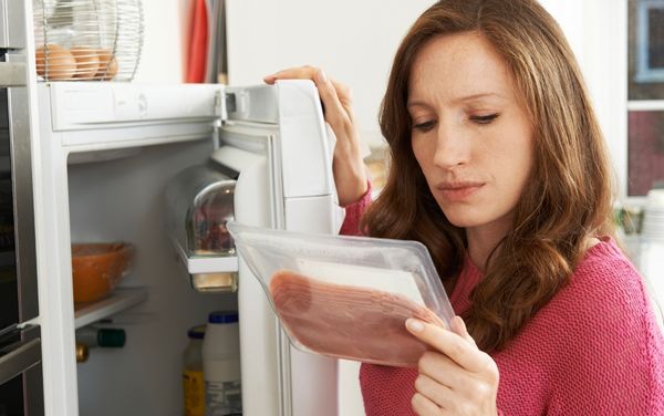 Woman checking the use-by date label on a packet of sliced meat in front of an open fridge