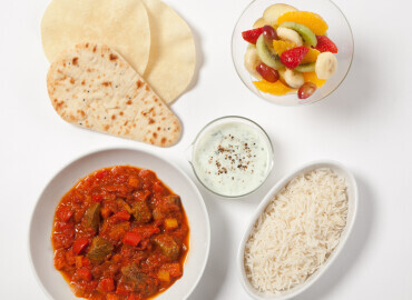 Photo of a beef curry with brown rice, raita, naan bread and poppadoms and a fruit salad