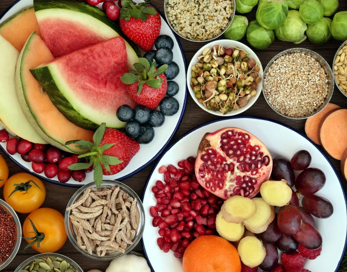Assorted fresh fruits and healthy grains displayed in various bowls and plates, including watermelon, strawberries, blueberries, pomegranate, oranges, and nuts.