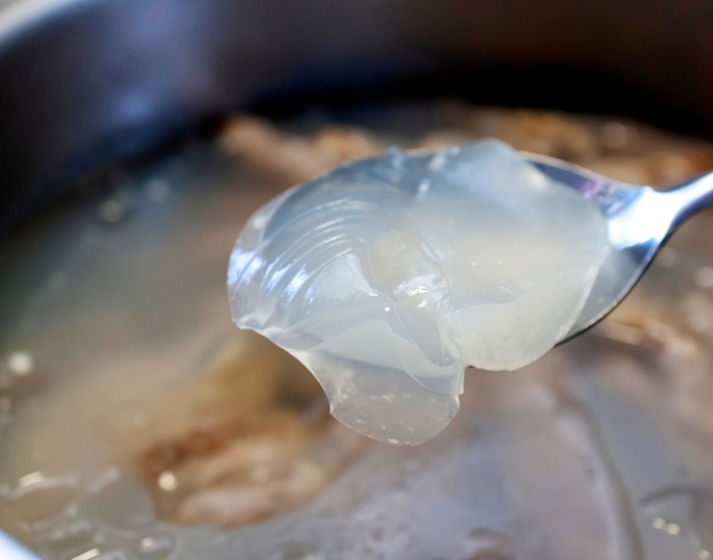 A close-up image of a spoon holding a translucent, gelatinous substance over a pot filled with a similar liquid