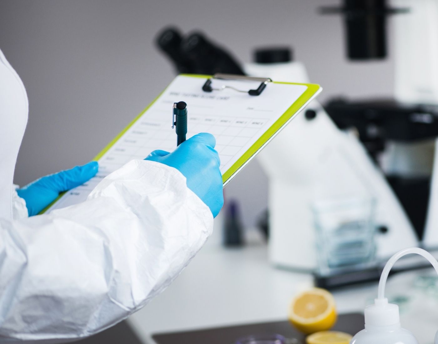 Person in blue gloves and a white lab coat holding a clipboard with a chart, standing in a laboratory with a microscope, containers, and a halved lemon in the background.