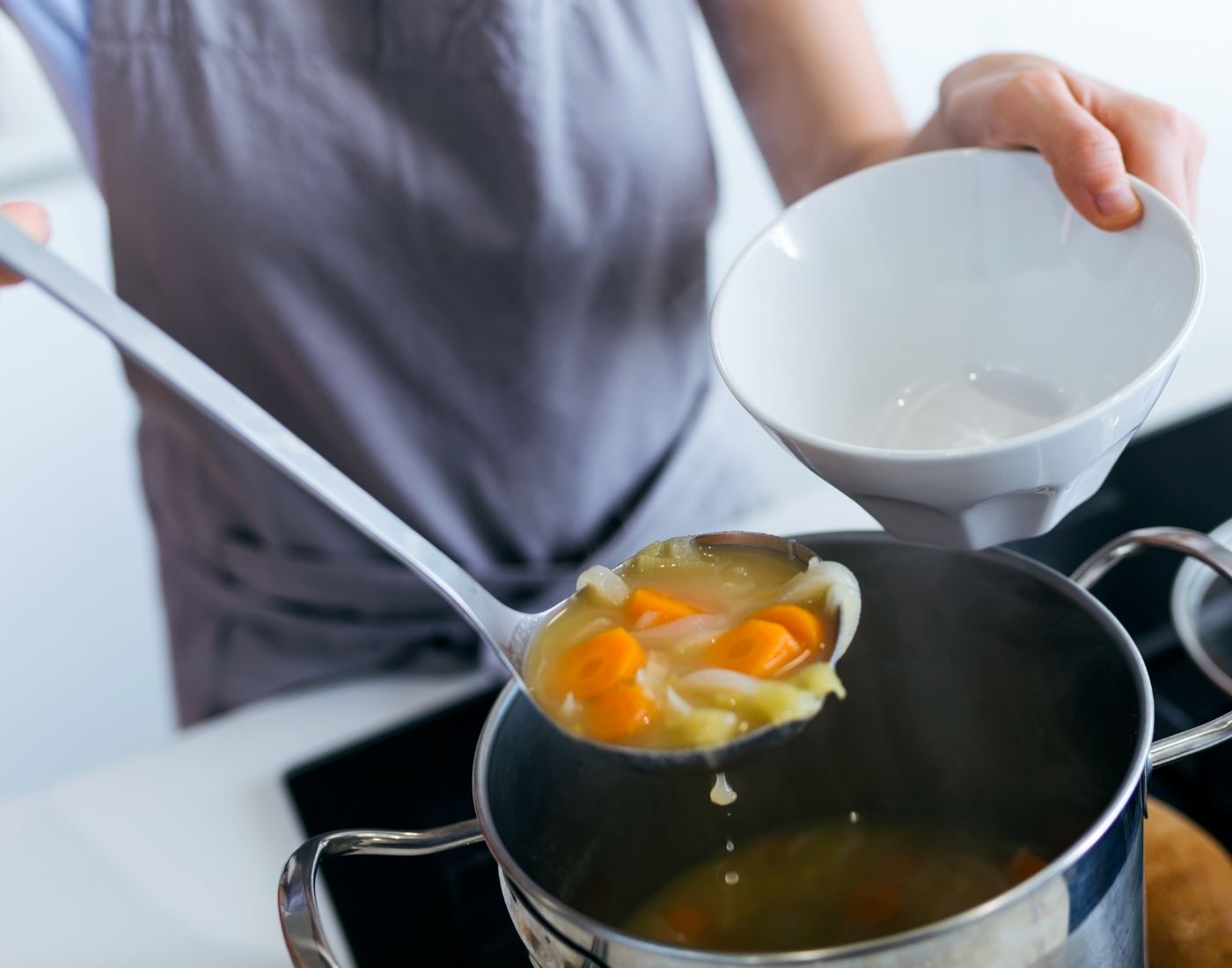 Person ladling homemade vegetable soup with carrots and celery from a pot into a white bowl in a kitchen setting.