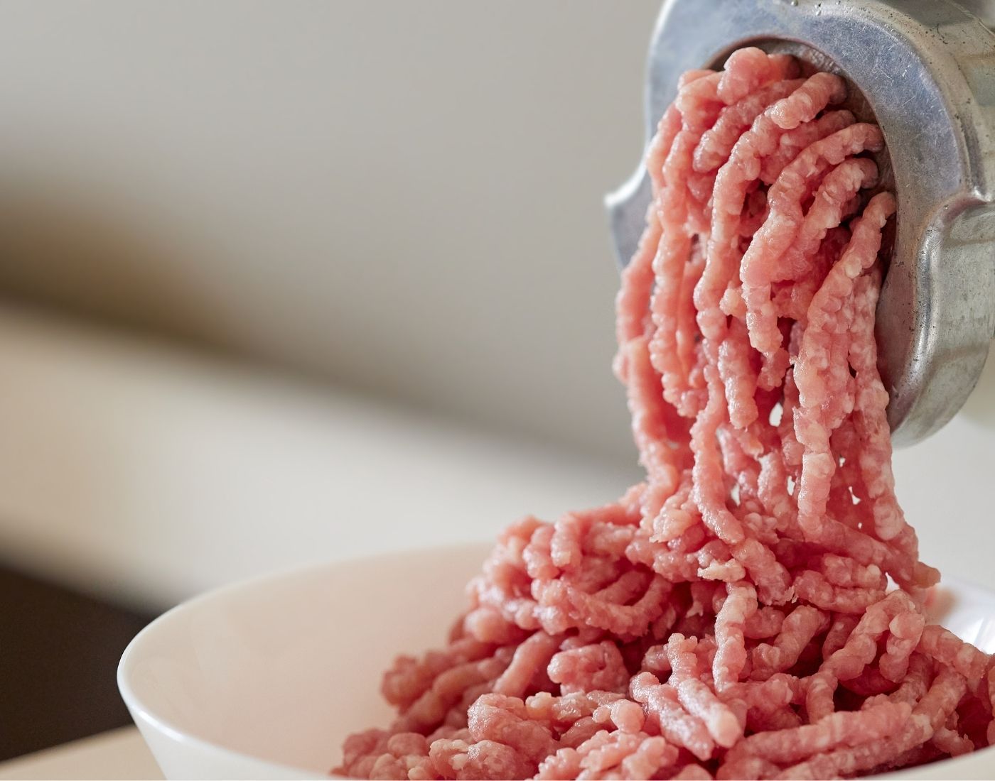 Ground meat being extruded from a metal meat grinder into a white bowl. The meat is pink and appears freshly ground.