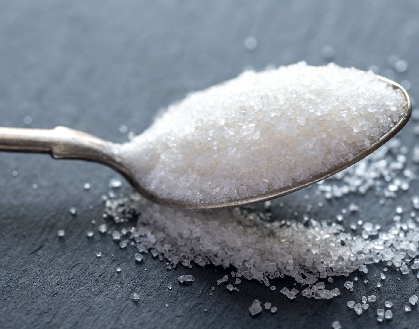 A close-up image of a spoon overflowing with granulated sugar, with some sugar grains scattered on a grey surface.