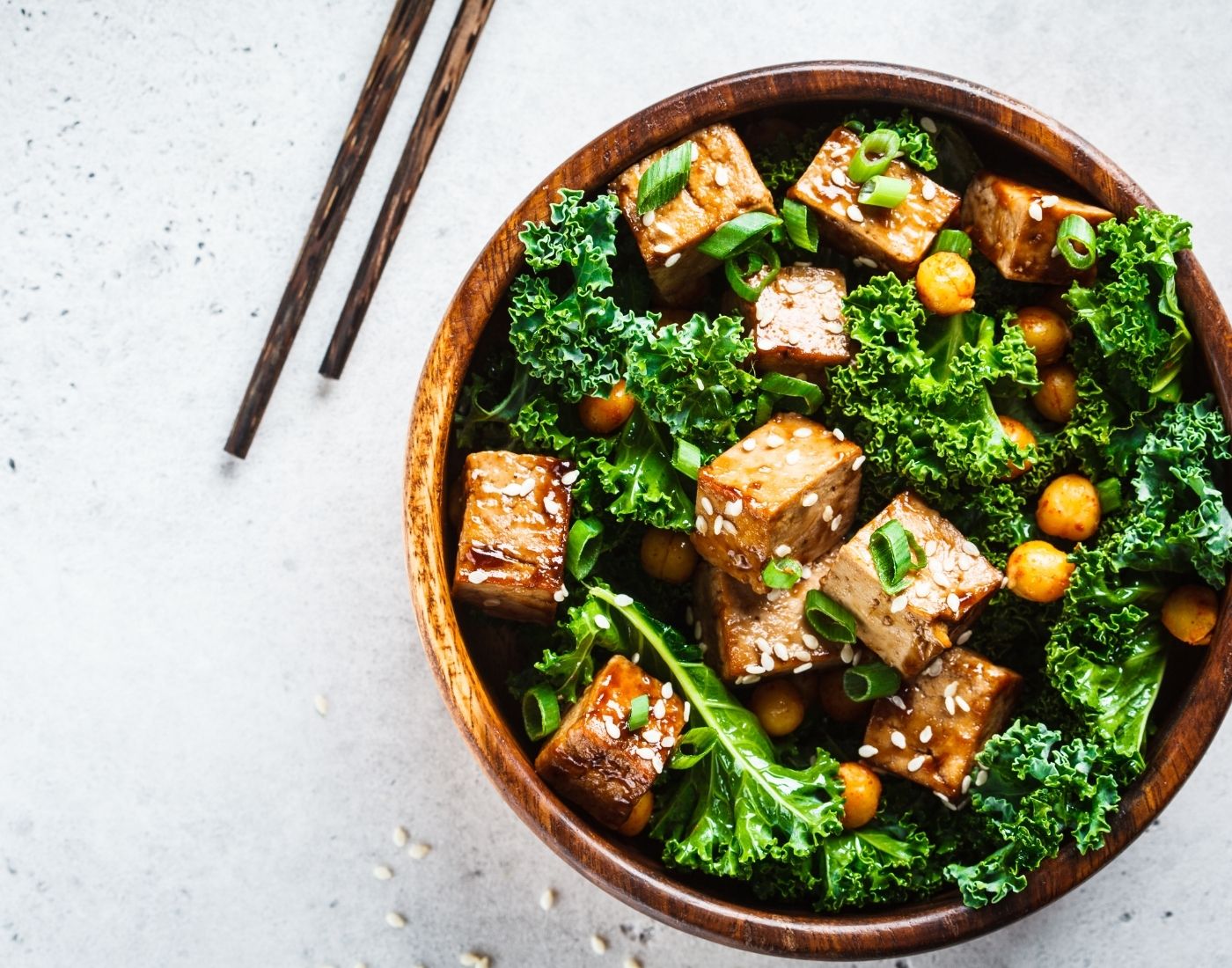 A bowl of tofu stir-fry with kale and chickpeas, garnished with sesame seeds and green onions, served with chopsticks on the side.