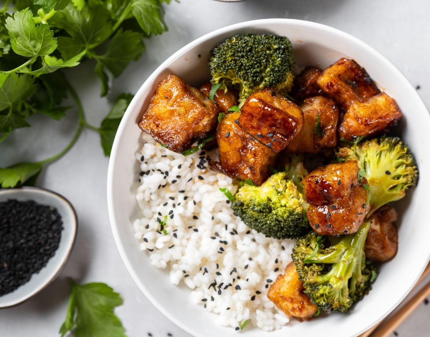 A bowl of white rice topped with black sesame seeds, crispy tofu cubes, and steamed broccoli florets, garnished with fresh parsley.