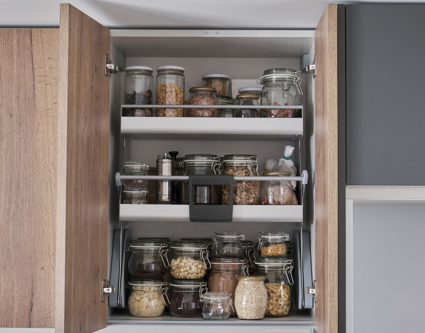 Open kitchen cabinet with three shelves containing glass jars filled with nuts, grains, spices, and other dry goods, each with metal or plastic lids.