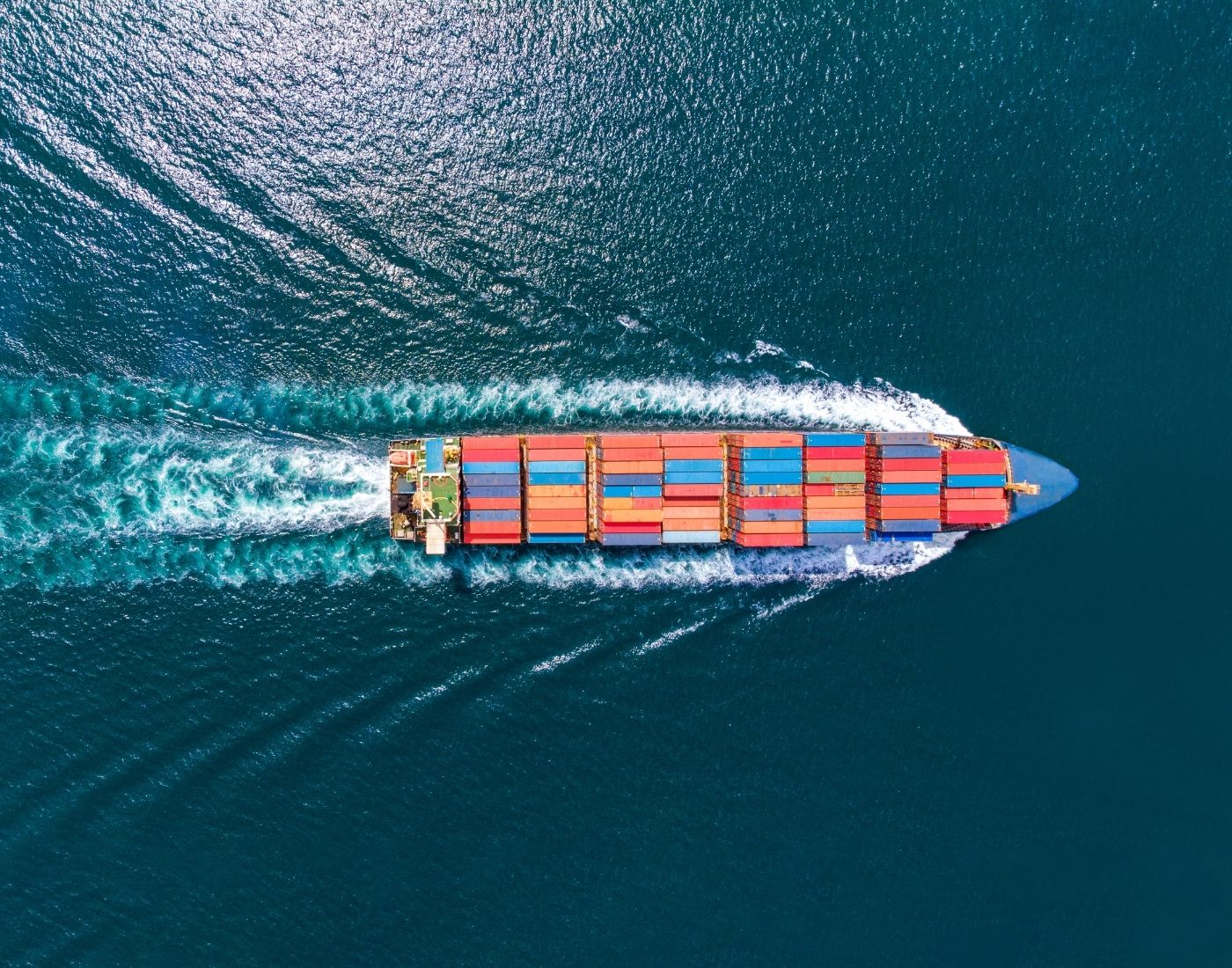 Aerial view of a large cargo ship carrying multiple coloured containers, sailing through blue ocean waters.