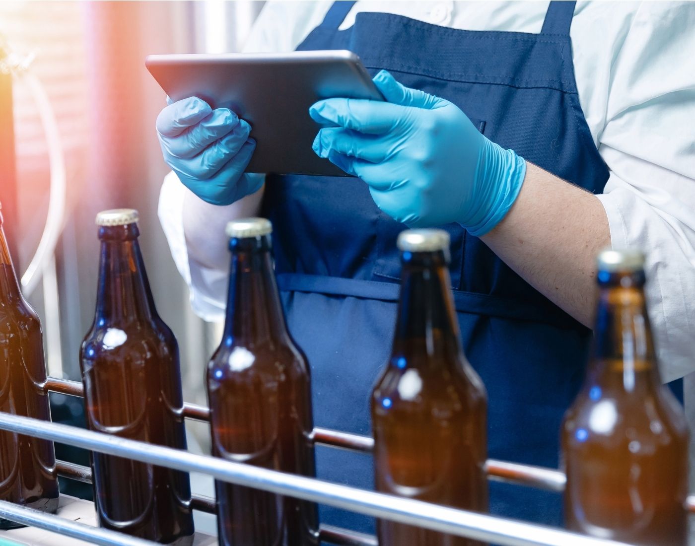 Four brown glass bottles on a production line. A worker is holding an iPad and checking them.