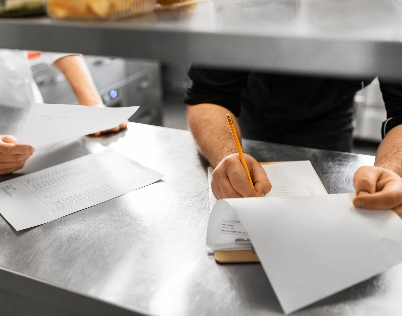 Two people's hands are seen holding paperwork and writing on a clip board in a restaurant kitchen serving area