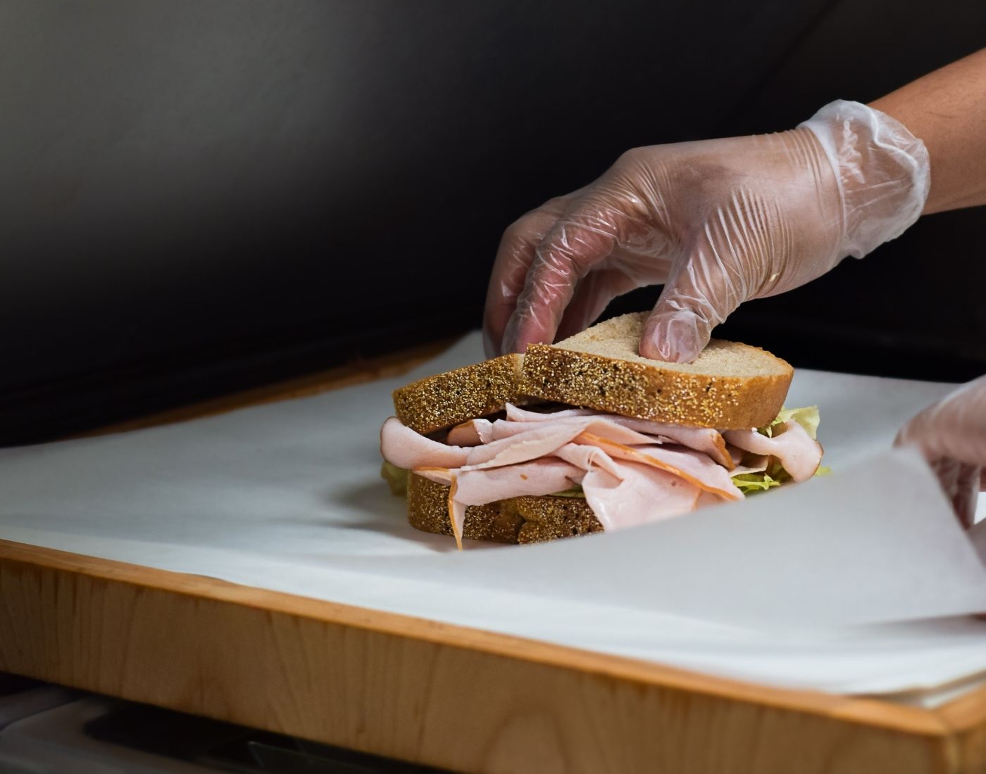 Hands in gloves preparing a sandwich with turkey and lettuce on whole grain bread on a kitchen counter.
