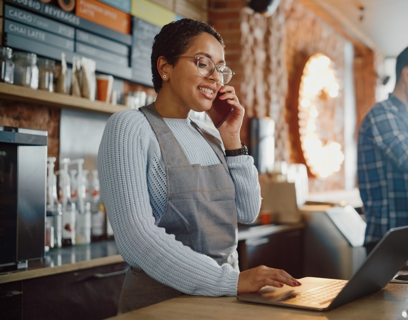 Cafe owner smiling while talking on the phone and working on her laptop.