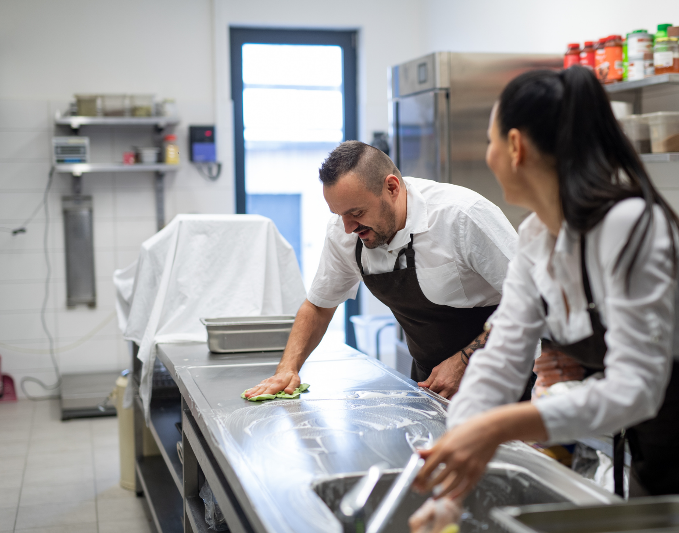 Male and female members of kitchen staff clean food preparation areas with soap and a cloth