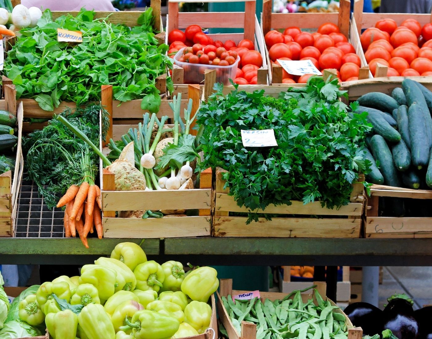 A market stall displays a colourful assortment of fresh vegetables in wooden crates, including leafy greens, tomatoes, cucumbers, carrots, bell peppers, and green beans, with handwritten labels marking types and prices.