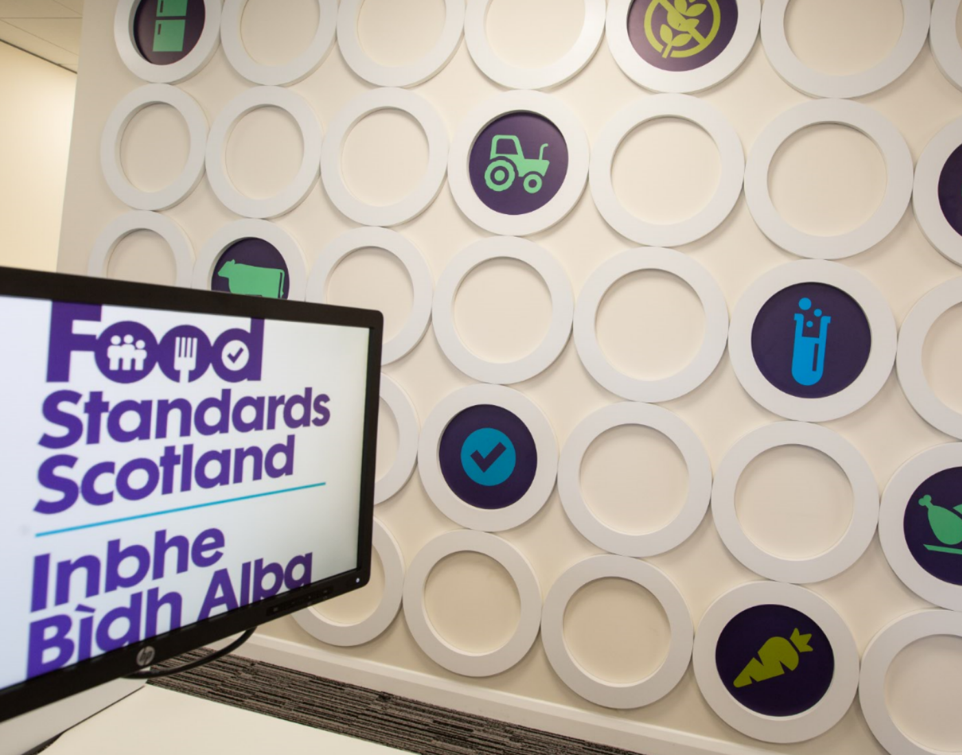 Inside of Food Standards Scotland's head office in Aberdeen, with a computer screen saying Food Standards Scotland in English and Gaelic, with a wall showing FSS related icons such as tractor, test tube and carrot. 