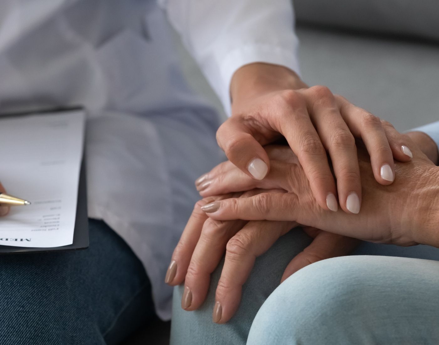 Close-up of a healthcare professional's hands comforting a patient's hands, with a clipboard visible in the background.
