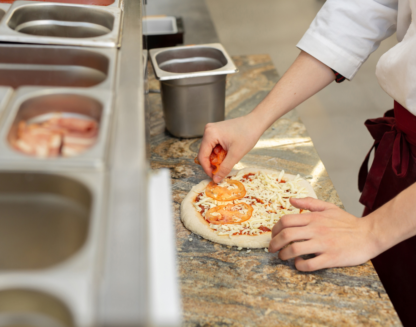 Persons hands are shown putting ingredients on a pizza in a restaurant kitchen