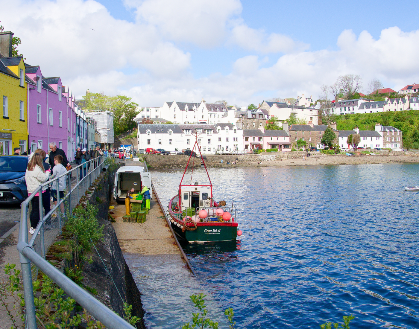 On a sunny day a fishing boat sits in calm water at Portree harbour. Fisherman are packing fish on the the harbour. Small businesses line the street nearby and some buildings of Portree are in the background.