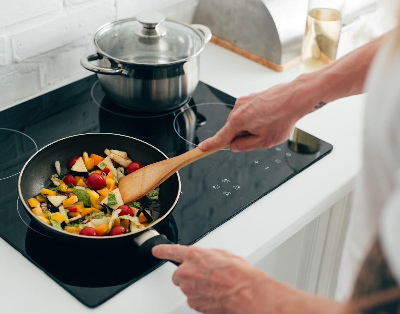 A person cooking vegetables in a frying pan on an induction stove, with a pot also on the stove in the background.