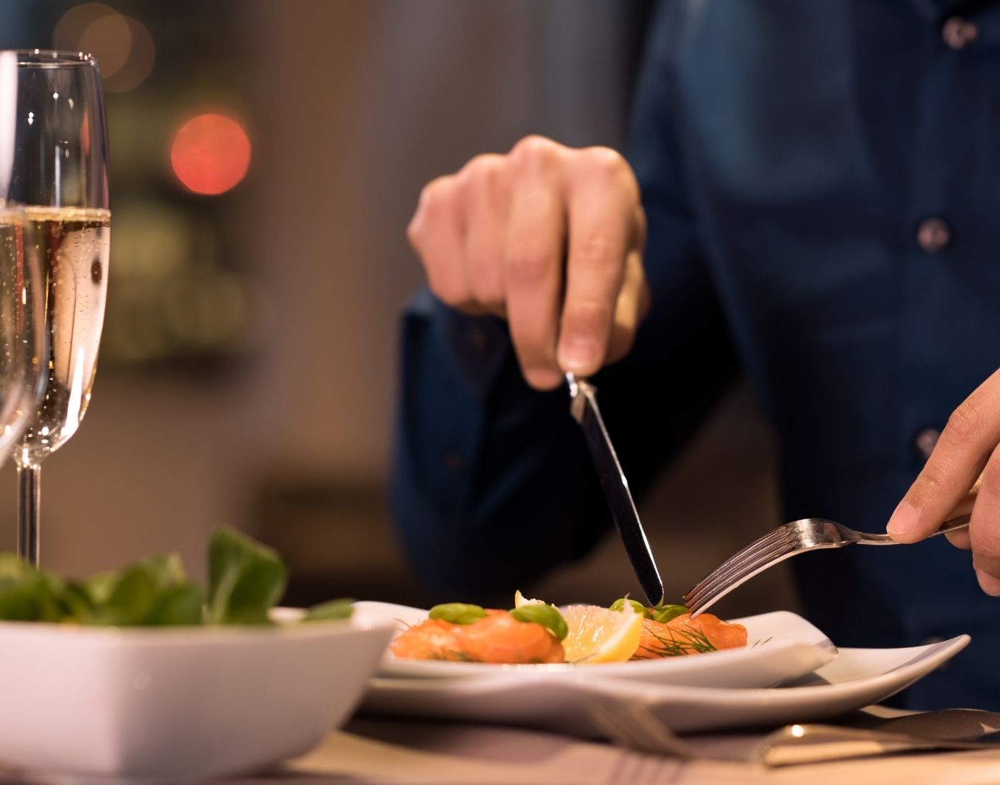 Close up of a man cutting up food on his plate. There's a glass of wine in front of him.