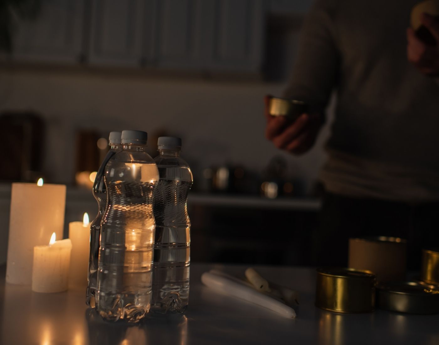 Dimly lit indoor scene with two bottles of water, several lit candles, a person holding a small can, additional cans on the table, and a flashlight or cylindrical object.