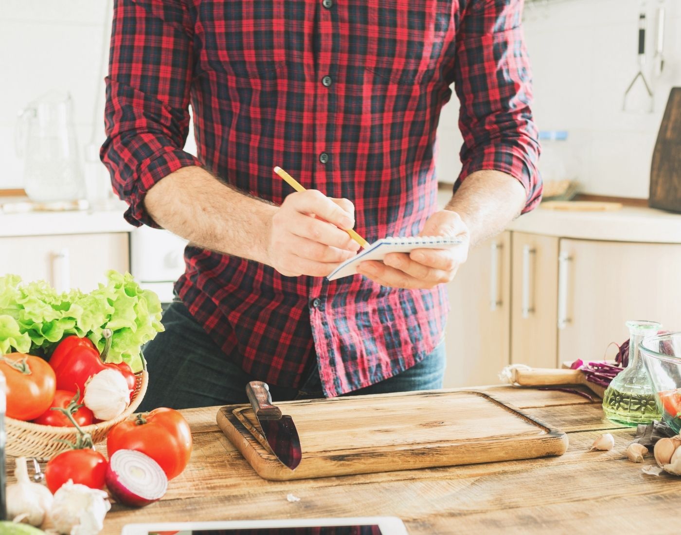 Person in a red and black checkered shirt standing in a kitchen, writing in a small notepad with a pencil. Various vegetables including tomatoes, lettuce, garlic, and onions are spread on the counter, along with a cutting board and knife.