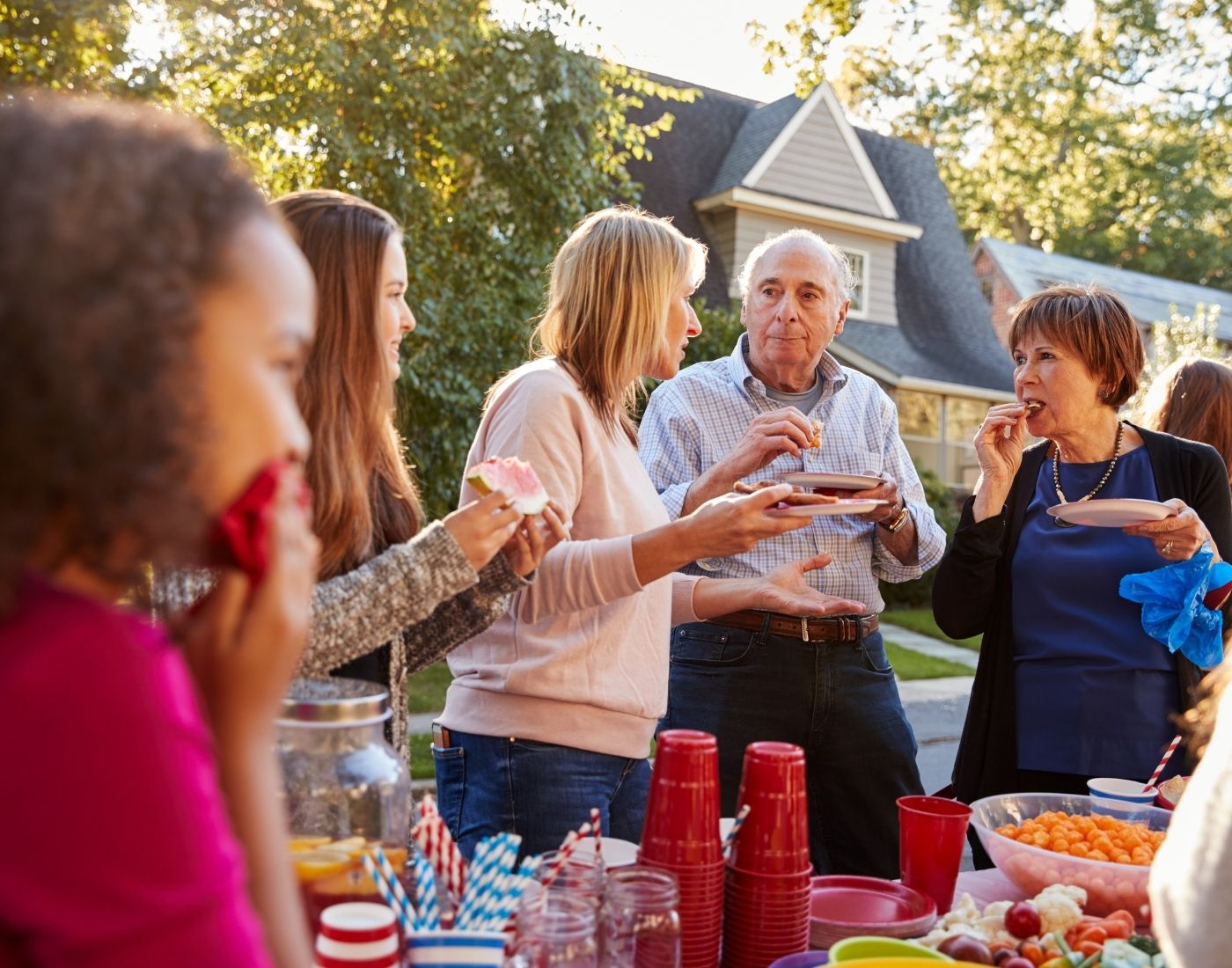 Group of people gathered outdoors around a table with food and red plastic cups, with trees and a house in the background. Faces are blurred for privacy
