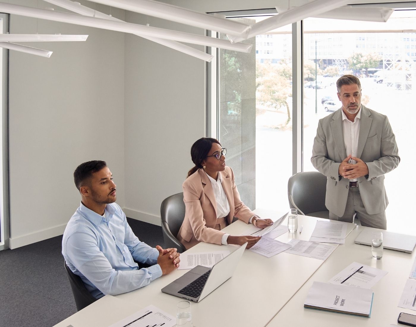 Three office workers in a meeting room, with one person standing and speaking while two others sit at a table with laptops and documents, in a bright, modern workspace.