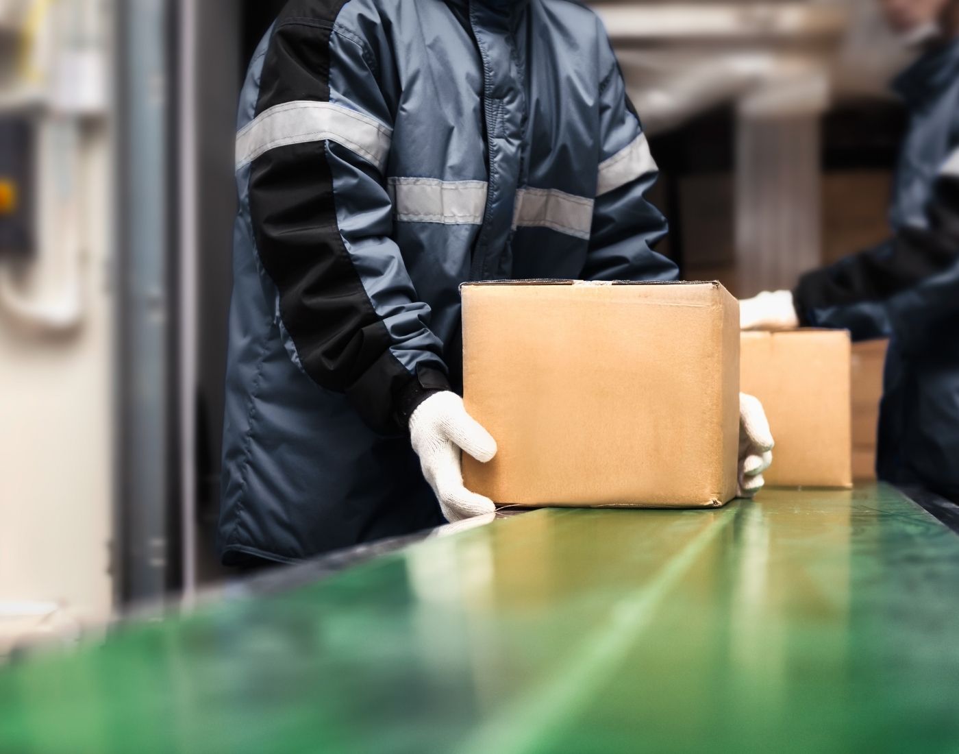Warehouse workers in blue reflective jackets and white gloves handling cardboard boxes on a green conveyor belt.