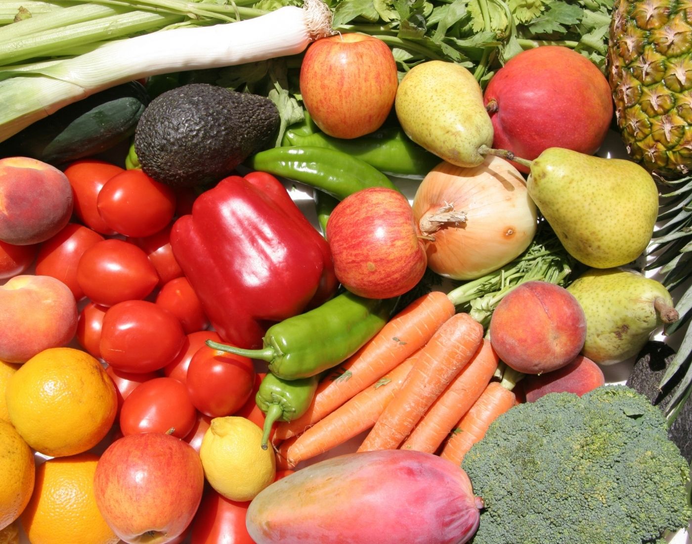 Assorted fresh fruits and vegetables including carrots, onions, a pineapple, mangoes, avocados, and a broccoli.