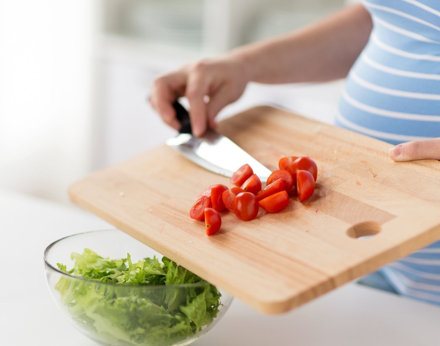 Pregnant woman with a chopping board and adding tomatoes to lettuce.