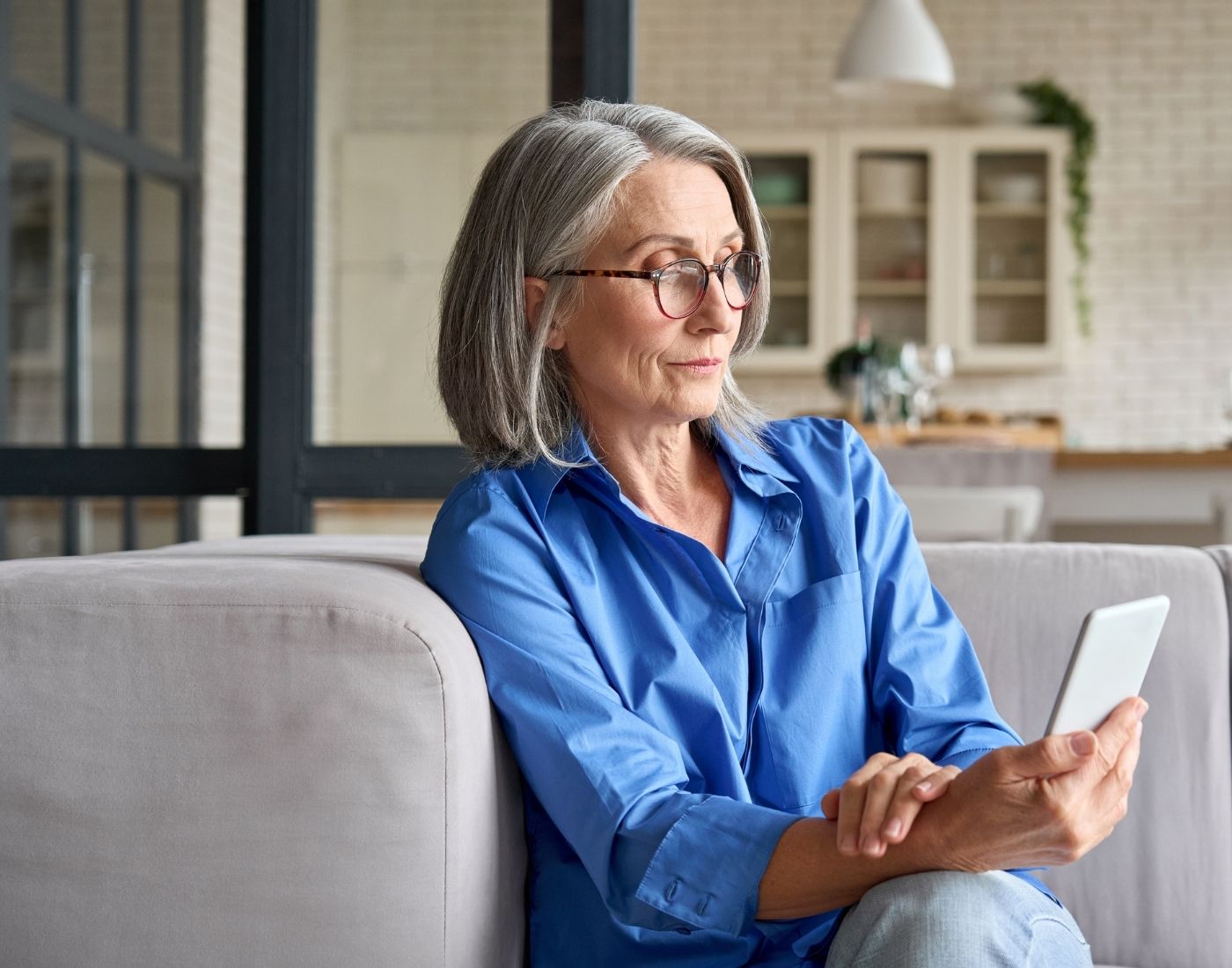 Person with gray hair sitting on a couch, wearing a blue shirt and holding a smartphone, with a modern kitchen featuring white cabinets and plants in the background.