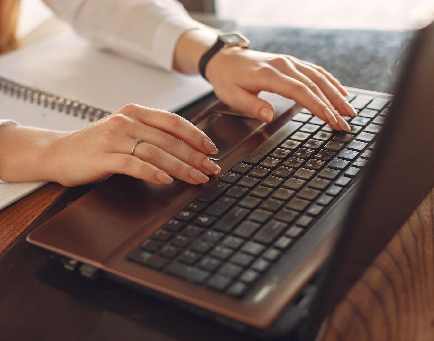 Person typing on a laptop keyboard with both hands, wearing a watch on the left wrist and a ring on the right hand, with a spiral-bound notebook open beside the laptop.