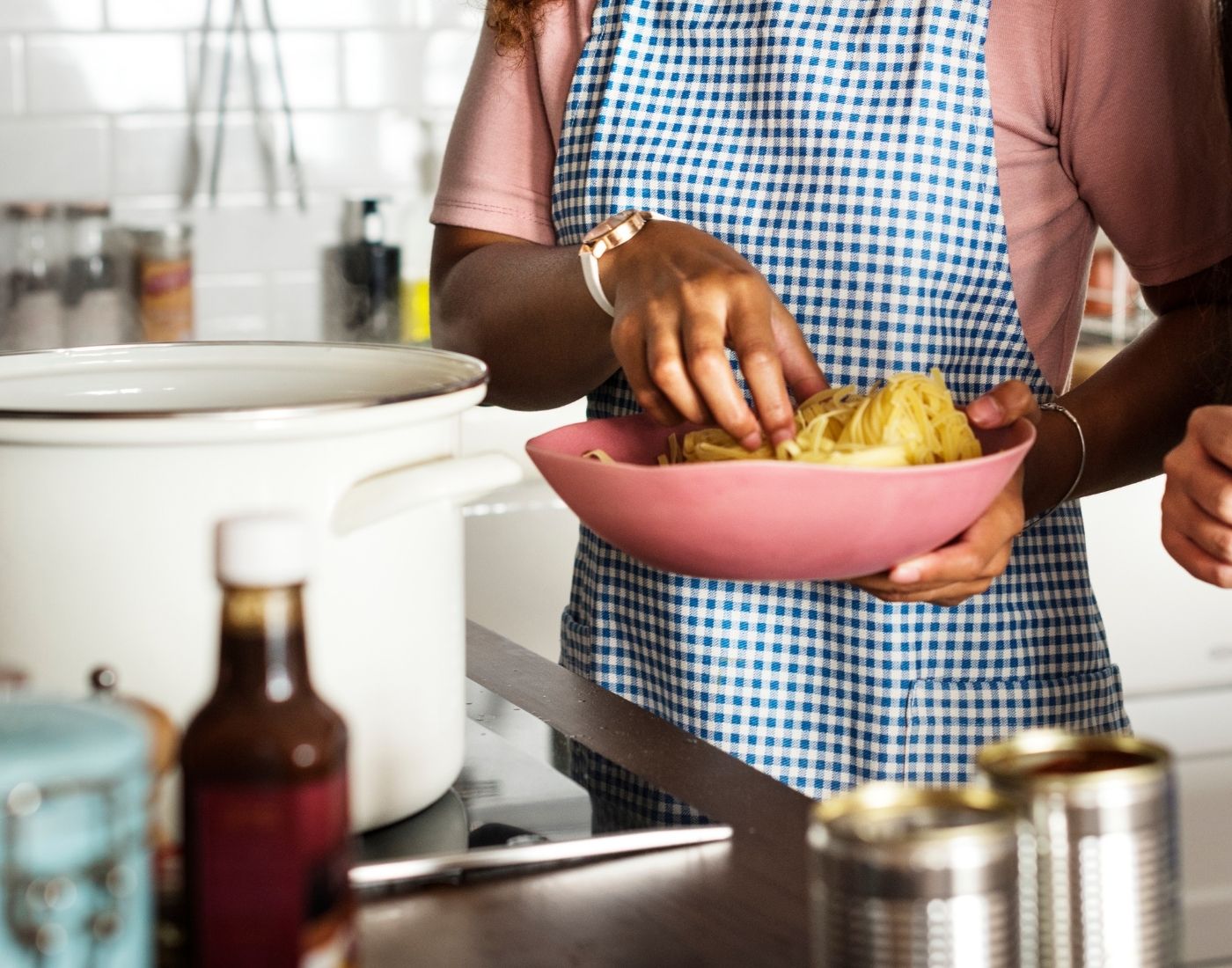 Person in a kitchen adding spaghetti from a pink pan into a pot, wearing a blue and white checkered apron. Ingredients and kitchenware are visible on the counter.