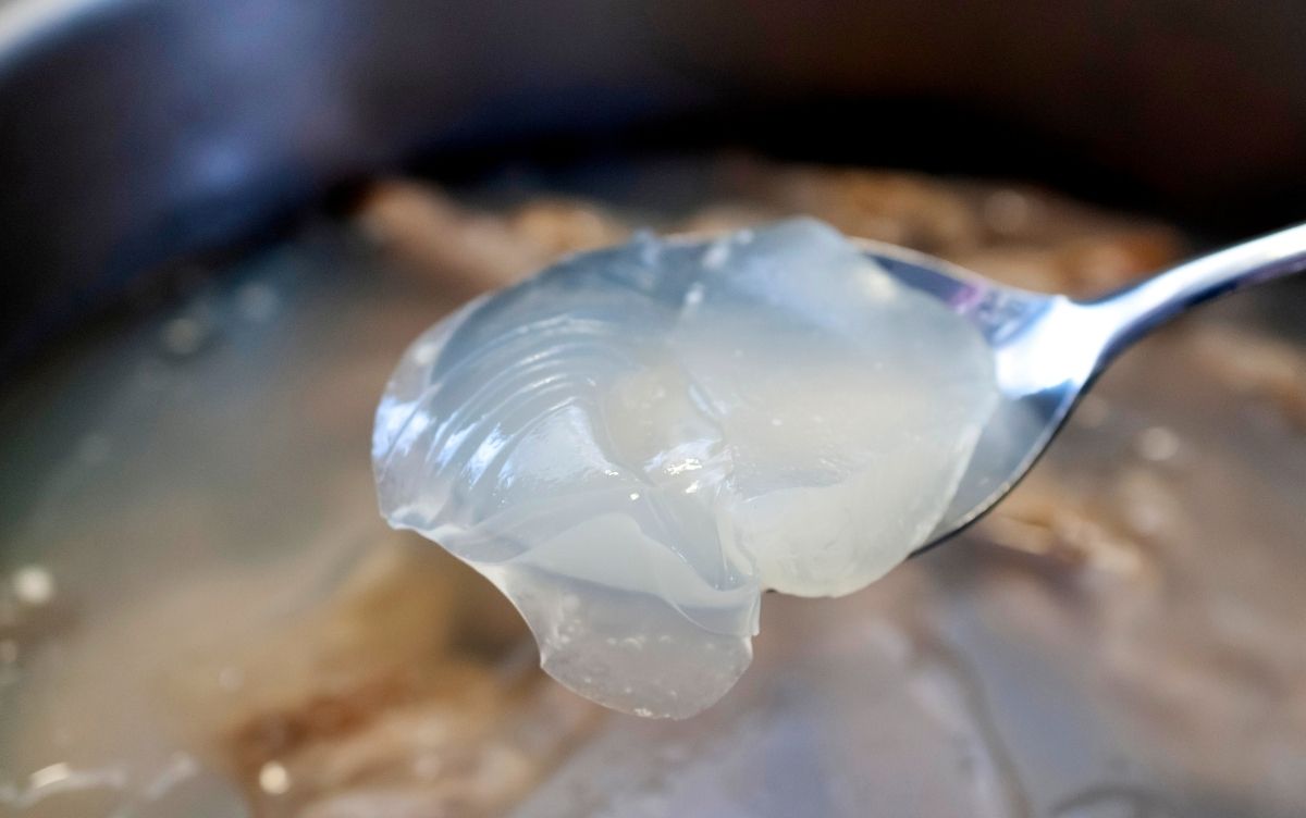 Close-up of a spoon holding translucent, gelatinous beef tendon and collagen, with a blurred pot of similar contents in the background.