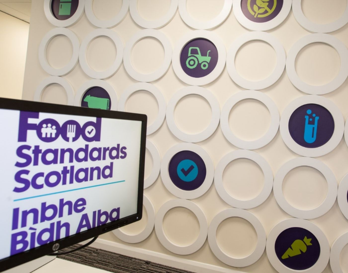 Wall with circular decorations featuring food and agriculture icons, alongside a computer monitor displaying the text 'Food Standards Scotland Inbhe Bìdh Alba'