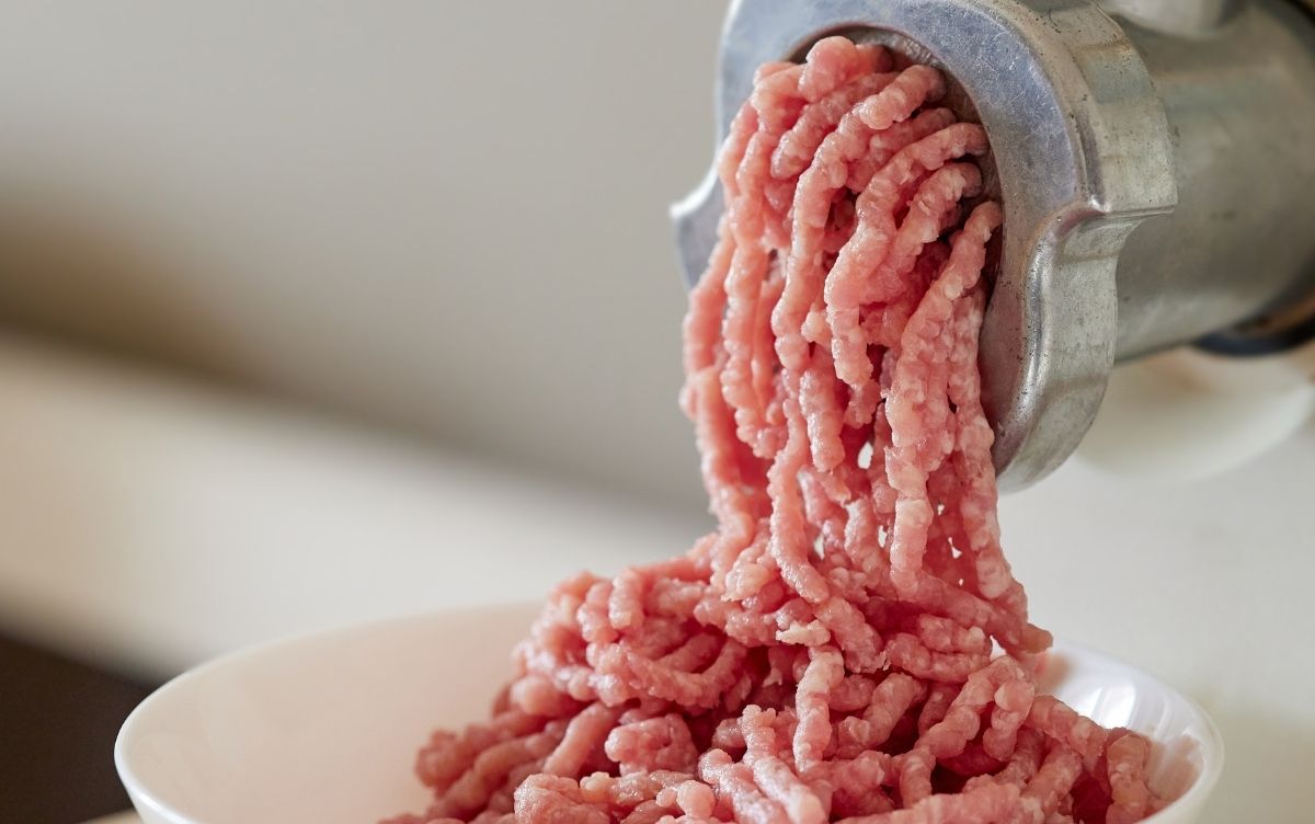 Ground meat being extruded from a metal meat grinder into a white bowl. The meat is pink and appears freshly ground.
