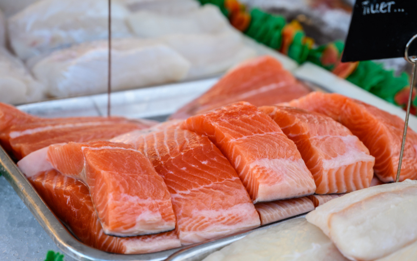 Fresh salmon fillets arranged on a metal tray at a seafood market, with bright orange flesh and white marbling. Other fish and a small sign are visible in the background.