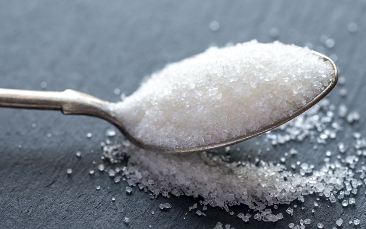 A close-up image of a spoon overflowing with granulated sugar, with some sugar grains scattered on a grey surface.