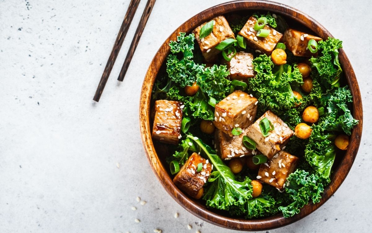 A bowl of tofu stir-fry with kale and chickpeas, garnished with sesame seeds and green onions, served with chopsticks on the side.