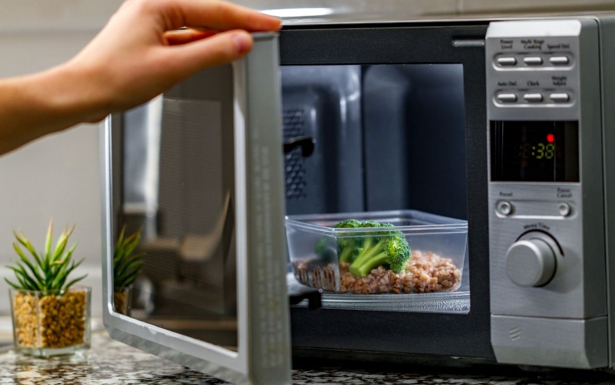 A person's hand opening a microwave containing a dish of broccoli and brown rice. The microwave digital display reads "0:38." A small potted plant is visible on the countertop.