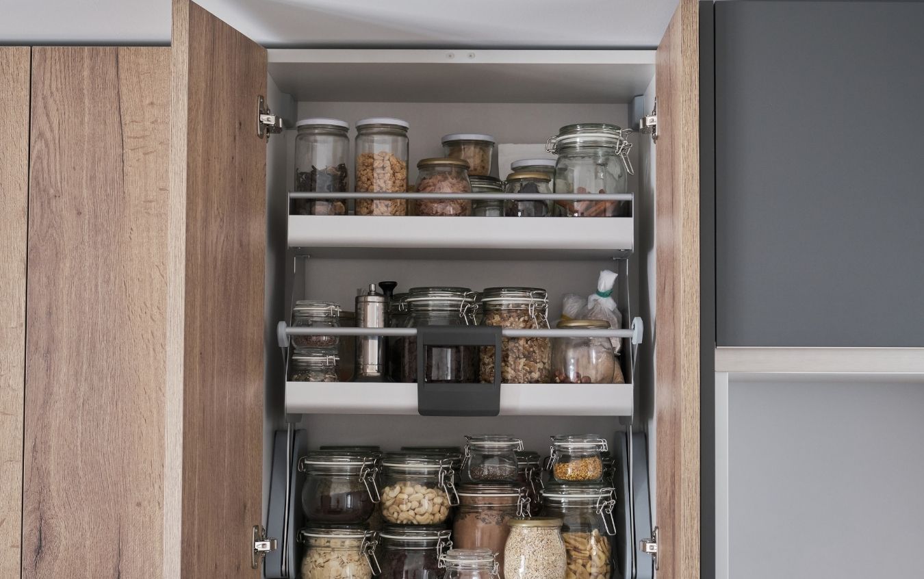 Open kitchen cabinet with three shelves containing glass jars filled with nuts, grains, spices, and other dry goods, each with metal or plastic lids