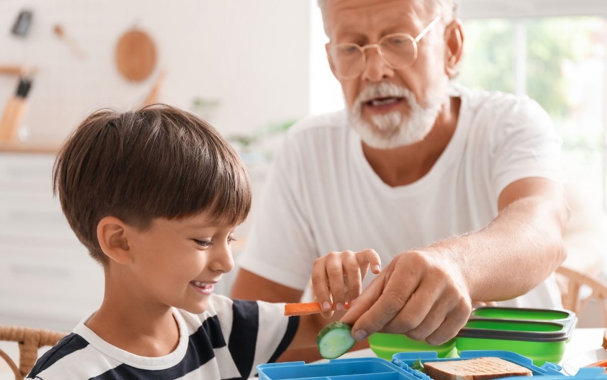 A primary school aged boy and his grandfather. They're preparing a packed lunch which includes cucumber slices and carrots.